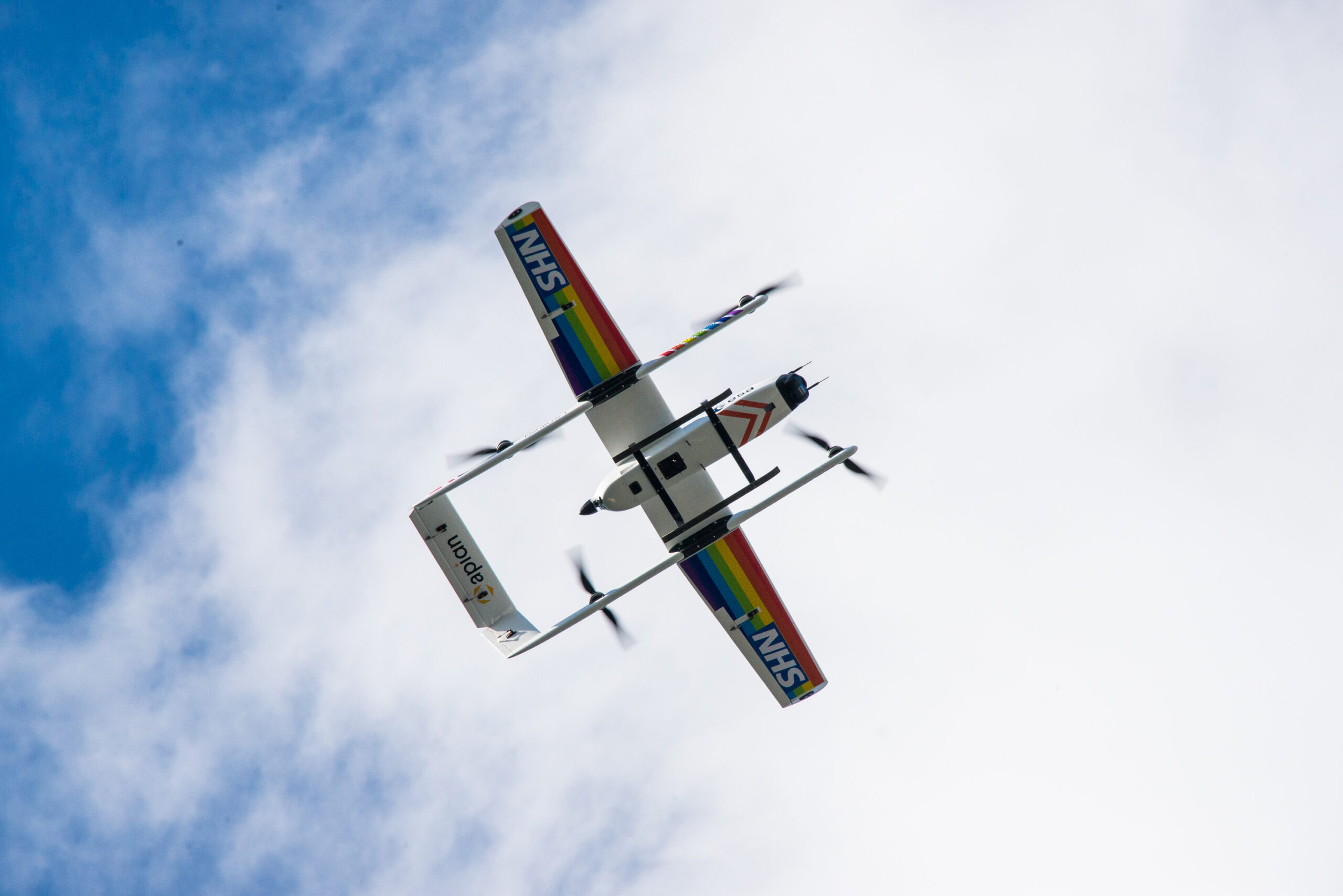 An Apian drone in flight, from below. The drone has NHS logos and rainbow flags on its wings.
